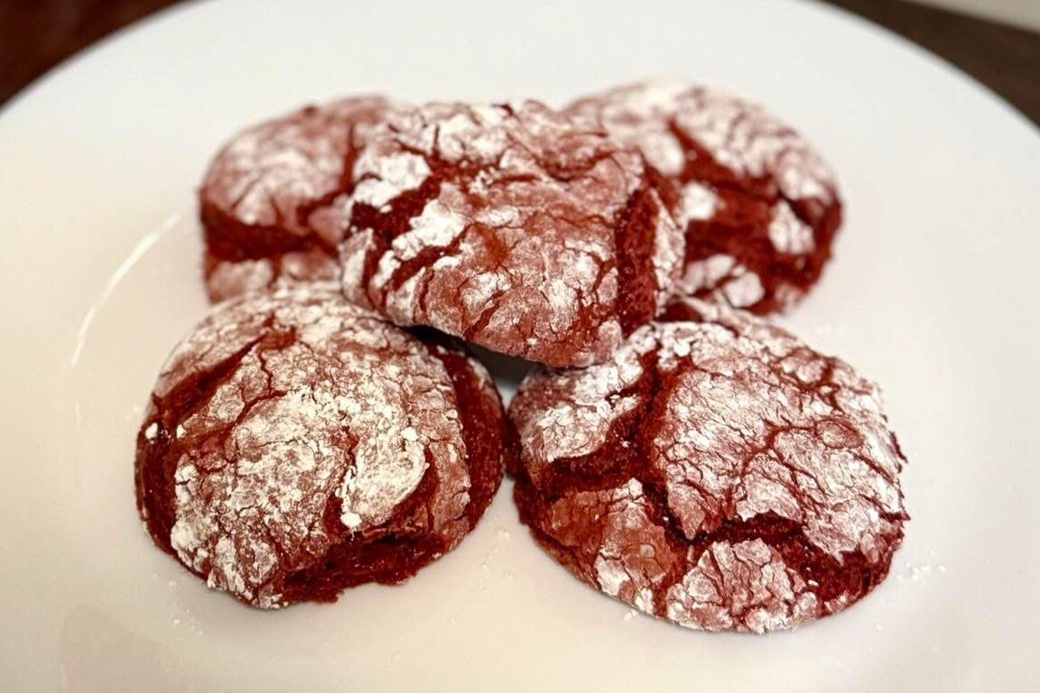 Galletas red velvet agrietadas con azúcar en polvo en un plato blanco, con una casita roja de cerámica al fondo