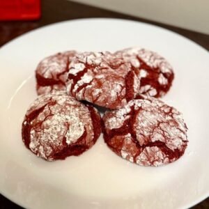 Galletas red velvet agrietadas con azúcar en polvo en un plato blanco, con una casita roja de cerámica al fondo
